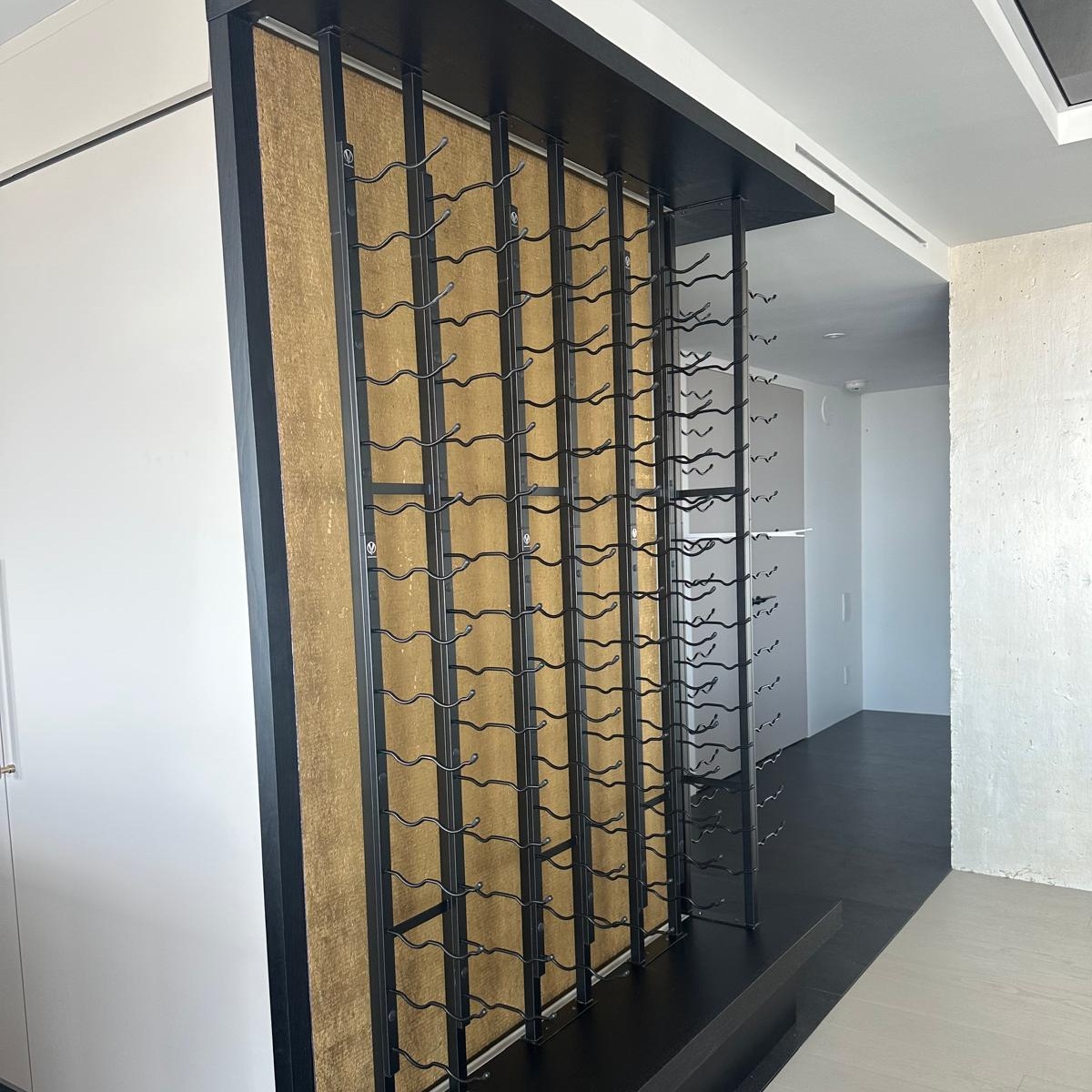 Muro de vino contemporáneo con soportes negros de pared para botellas instalados sobre un panel decorativo dorado.
