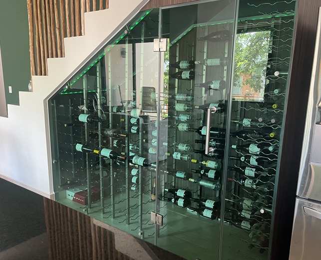 Cava de vino acristalada bajo una escalera equipada con soportes de pared negros para botellas de vino y presentación con las etiquetas visibles.
